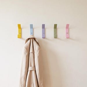 May include: Five wall-mounted metal coat hooks in yellow, light blue, lavender, sage green, and pink, each secured with a single screw. A beige trench coat hangs from the light blue hook, displaying a collar, lapels, and a belted waist detail.