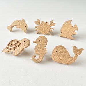 Könnte beinhalten: Ein Set aus sechs hellen Holzhaken in Form von Meerestieren, darunter ein Delfin, eine Krabbe, ein Fisch, eine Meeresschildkröte, ein Seepferdchen und ein Wal. Jedes Stück zeigt eine natürliche Holzmaserung und ein kleines kreisförmiges Loch.