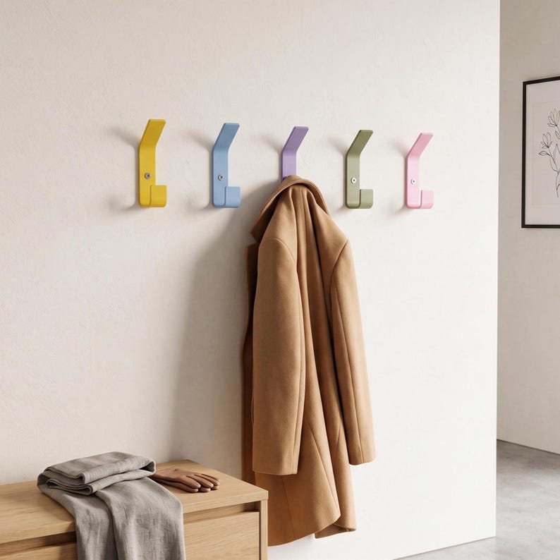 May include: Five wall-mounted metal hooks in yellow, light blue, purple, olive green, and pink, each secured with a single visible screw. A tan coat hangs from the purple hook, while a folded gray fabric and brown gloves rest on a wooden surface.