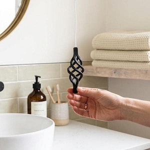 Puede incluir: Tirador de luz de baño negro y decorativo con un diseño retorcido. El tirador está suspendido por un cordón blanco. El fondo incluye un lavabo, un espejo y toallas.