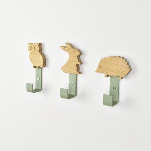 Może przedstawiać: Trzy haczyki ścienne z jasnobrązowymi drewnianymi sylwetkami sowy, królika i jeża. Każda drewniana figurka jest przymocowana do szałwiowo-zielonej metalowej podstawy za pomocą pojedynczej srebrnej śruby, przeznaczonej do montażu na płaskiej powierzchni.