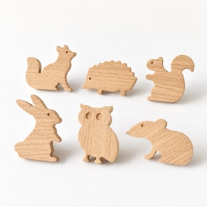 May include: A set of six light-coloured wooden animal-shaped knobs. The collection includes silhouettes of a fox, a hedgehog, a squirrel, a rabbit, an owl, and a mouse, each featuring a smooth, natural wood grain finish and a small circular eye detail.