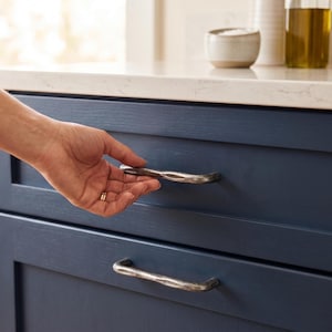 May include: A hand opening a navy blue cabinet drawer with a textured, silver-toned handle. The drawer is part of a kitchen cabinet set. The cabinet has a white countertop and other kitchen items in the background.