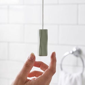 Könnte beinhalten: Ein salbeigrüner Lichtzugschalter mit vertikaler Wellenstruktur, befestigt an einer weißen geflochtenen Schnur. Das zylindrische Objekt hat eine glatte, matte Oberfläche und eine markante, konturierte Form zur Bedienung von Deckenleuchten.