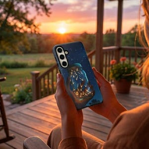 Op de afbeelding: Een smartphonehoesje met een donkerblauwe achtergrond met een pot gevuld met gloeiende lichten. De hoes heeft een sterrenhemelontwerp en wordt vastgehouden door een persoon op een houten veranda met een zonsondergang op de achtergrond.