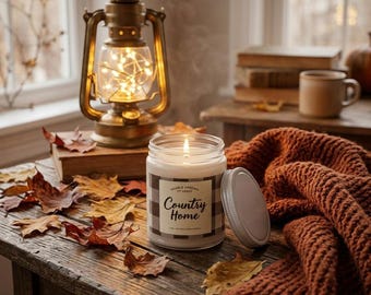 May include: A lit candle in a clear glass jar with a white label featuring a black and white buffalo check border and the text Country Home. The candle sits on a wooden surface with scattered brown autumn leaves and a rust-colored knit blanket.