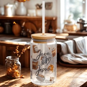 Peut inclure: Un gobelet en verre transparent avec un couvercle en bambou et une paille transparente. Le gobelet porte les mots "Java & Jesus" en écriture cursive, ainsi que des illustrations sur le thème du café. Le gobelet est posé sur une surface en bois.