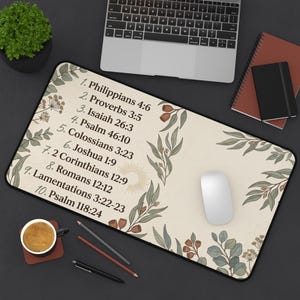 May include: A rectangular desk mat with a cream background featuring a list of ten Bible verses in black text, surrounded by botanical illustrations of green leaves and brown berries, with a sunburst icon near the center of the list.