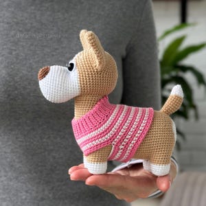 Op de afbeelding: Een gehaakt hondenspeeltje van beige en wit garen, met een bruine neus, zwarte ogen en een roze met wit gestreepte trui. Het kleine knuffeldier heeft puntige oren, een korte staart en witte pootjes, met de tekst MommyPatterns zichtbaar.