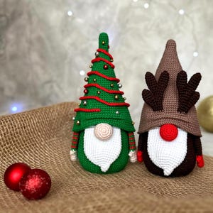 May include: Two handmade crochet Christmas gnomes. One is green with red accents and gold beads, resembling a Christmas tree. The other is brown with reindeer antlers and a red nose. Red Christmas ornaments are in the foreground.