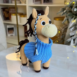 Puede incluir: Figura de caballo de ganchillo con cuerpo marrón claro, hocico blanco y suéter azul. El caballo tiene pelo castaño oscuro y un acento trenzado. Los ojos son grandes y negros. El caballo está sobre una superficie blanca.