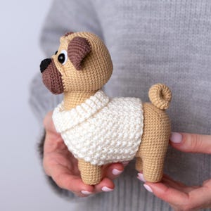May include: A crocheted pug dog figurine in tan, brown, and cream colors. The pug has a cream-colored sweater and is held in a person's hands. The dog has a short tail and a distinctive pug face.