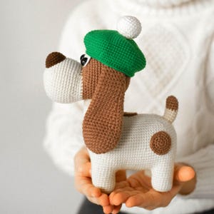 Pode incluir: Um brinquedo de cachorro de crochê com corpo branco, orelhas marrons e boina verde com pompom branco. O cachorro tem uma mancha marrom e uma cauda pequena. O brinquedo é segurado nas mãos.