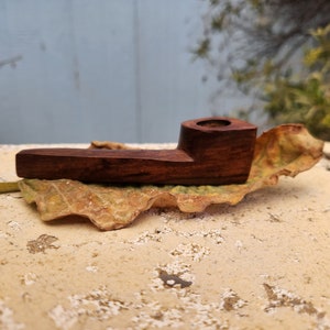 Puede incluir: Una pequeña pipa de madera marrón oscuro que descansa sobre una hoja seca. La pipa tiene un diseño sencillo con una cazoleta redonda y una boquilla ligeramente curvada.