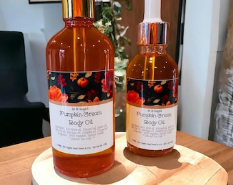 May include: Two amber glass bottles with gold lids and droppers filled with pumpkin cream body oil. The bottles have labels with the text "Be In Magick Pumpkin Cream Body Oil" and a fall-themed design with pumpkins, leaves, and flowers. The bottles are sitting on a wooden surface.