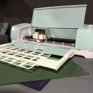 May include: A light green and white electronic cutting machine with the lid open. The machine has a cutting mat and a gridded cutting surface.