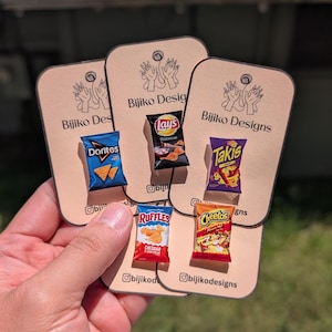Peut inclure: Cinq épinglettes miniatures en forme de sachets de snacks représentant Doritos, Lay's Barbecue, Takis, Ruffles Cheddar & Sour Cream et Cheetos. Chaque épinglette est fixée sur une carte beige clair marquée Bijiko Designs avec un dessin de mains.