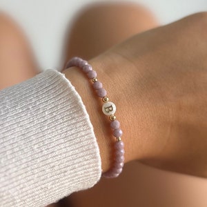 Puede incluir: Un brazalete delicado con pequeñas cuentas de color morado claro y un dije de oro con la letra "B" sobre un fondo blanco.