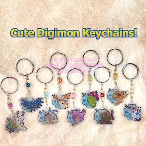 May include: A collection of Digimon keychains with various character designs. Each keychain features a clear acrylic charm with a colorful illustration, attached to a silver keyring with decorative beads. The text "Cute Digimon Keychains!" is displayed at the top.