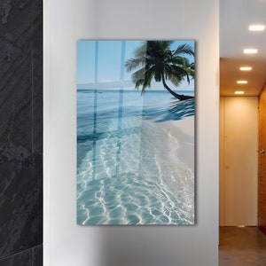 Peut inclure: Une photographie d'une scène de plage tropicale avec un palmier sur le rivage et une eau bleu clair. L'eau est peu profonde et le sable est blanc. L'image est encadrée et accrochée à un mur dans un couloir.