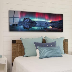 Pode incluir: Uma impressão retangular de arte de parede que retrata uma vibrante aurora boreal sobre uma paisagem montanhosa. A obra apresenta tons de vermelho, verde e azul, criando uma cena dramática e colorida. A impressão é montada acima de uma cama com almofadas.