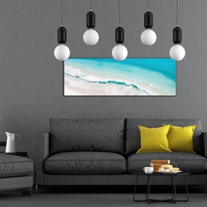 May include: A gray sofa with two yellow pillows in front of a gray wall with a framed photograph of an aerial view of a beach with turquoise water and white sand. There are six black and white pendant lights hanging from the ceiling.