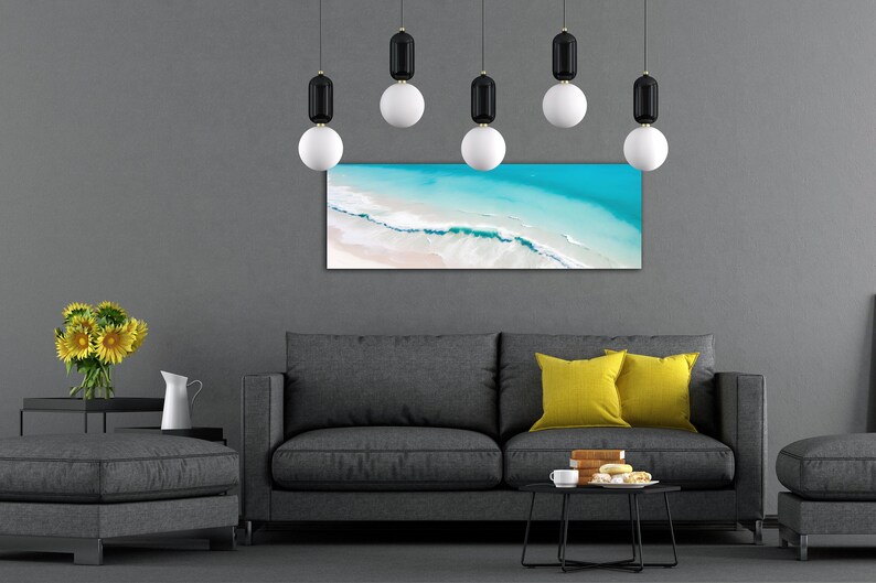 May include: A gray sofa with two yellow pillows in front of a gray wall with a framed photograph of an aerial view of a beach with turquoise water and white sand. There are six black and white pendant lights hanging from the ceiling.