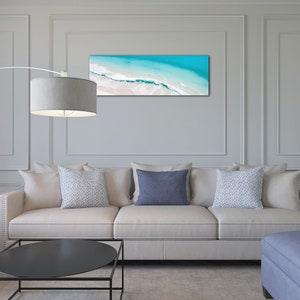 May include: A white couch with three pillows, two patterned and one solid blue, sits in a living room with a black coffee table and a blue ottoman. A floor lamp with a gray shade is in the corner of the room. A framed photograph of an aerial view of a beach with turquoise water and white sand hangs on the wall.