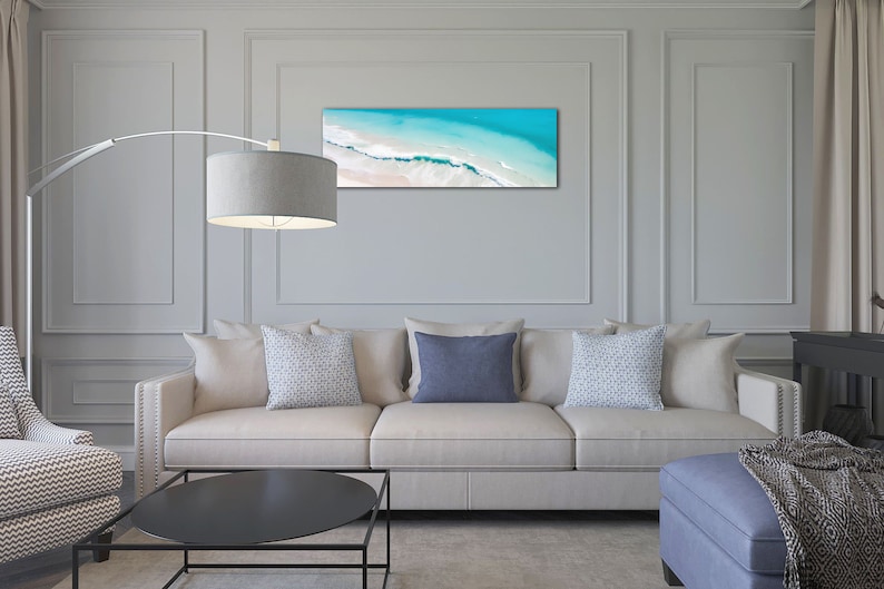 May include: A white couch with three pillows, two patterned and one solid blue, sits in a living room with a black coffee table and a blue ottoman. A floor lamp with a gray shade is in the corner of the room. A framed photograph of an aerial view of a beach with turquoise water and white sand hangs on the wall.