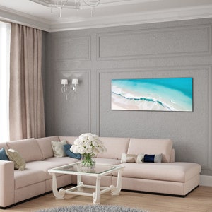 May include: A light beige sectional sofa with pillows in a living room setting. The sofa is in front of a gray wall with a framed print of an aerial view of a beach with turquoise water and white sand.