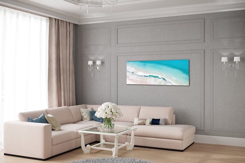 May include: A light beige sectional sofa with pillows in a living room setting. The sofa is in front of a gray wall with a framed print of an aerial view of a beach with turquoise water and white sand.