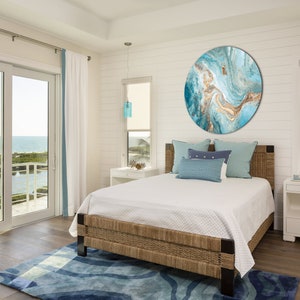 May include: A bedroom with a large, round, blue and gold abstract painting on the wall. The bed has a white duvet and blue pillows. The room has a large window with a view of the ocean.