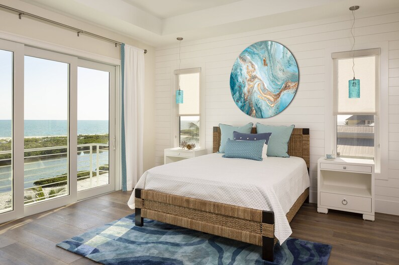 May include: A bedroom with a large, round, blue and gold abstract painting on the wall. The bed has a white duvet and blue pillows. The room has a large window with a view of the ocean.