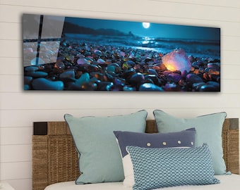 Puede incluir: Una impresión de arte mural panorámica que representa una escena de playa a la luz de la luna. La obra de arte presenta un primer plano de piedras y guijarros coloridos y brillantes en la orilla, con el océano y la luna llena en el fondo. La impresión se muestra encima de una cama con almohadas.