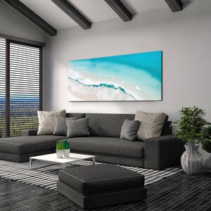 May include: A gray sectional sofa with pillows sits in a living room with a large abstract painting of a beach scene above it. The painting features blue water, white sand, and white waves. A small coffee table sits in front of the sofa, and a black and white rug is on the floor.
