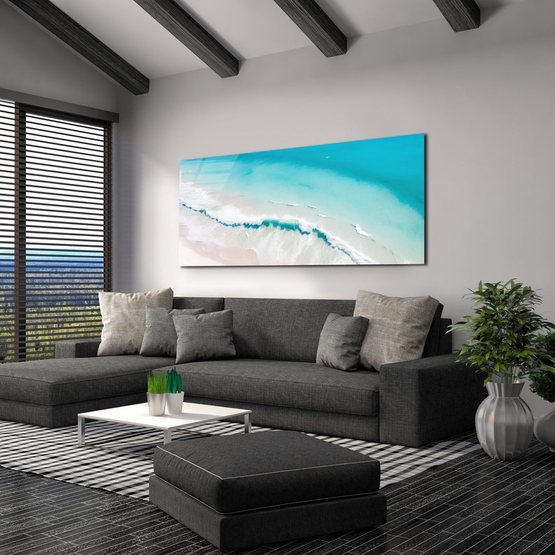 May include: A gray sectional sofa with pillows sits in a living room with a large abstract painting of a beach scene above it. The painting features blue water, white sand, and white waves. A small coffee table sits in front of the sofa, and a black and white rug is on the floor.