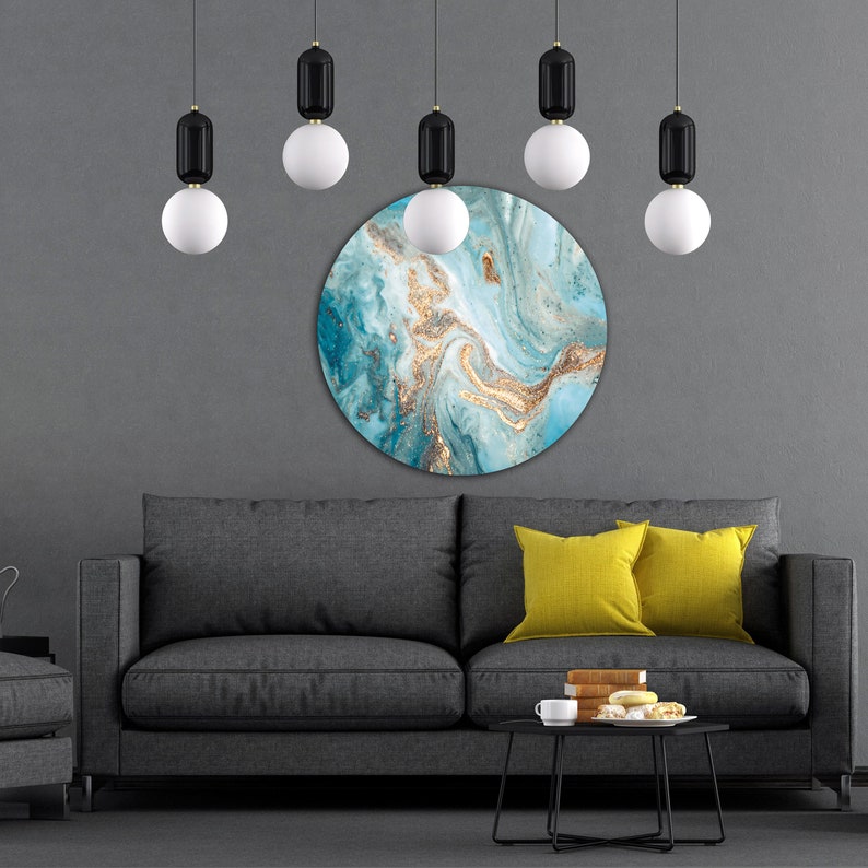 May include: A round, blue and gold abstract wall art print with a swirling pattern. The print is hanging on a gray wall above a gray sofa with yellow throw pillows.