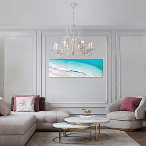 May include: A white crystal chandelier with a white metal frame hangs from the ceiling in a living room. The chandelier has multiple crystal drops and is lit with white bulbs. The room has a white couch, two white coffee tables, and a white armchair with a pink pillow.