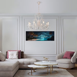 May include: A white crystal chandelier with a white metal frame hangs from the ceiling in a living room. The chandelier has eight lights and is surrounded by white walls and a white sofa. A tropical beach scene is hanging on the wall behind the chandelier.