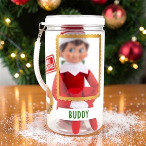 May include: A clear plastic cylindrical container with a white screw-top lid and a white coiled wrist strap. The container features a gold-bordered frame containing a doll, a red label reading HANDLE WITH CARE, and the name BUDDY in green.