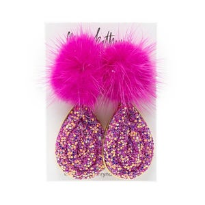 Peut inclure: Une paire de boucles d'oreilles fuchsia vives. Chaque boucle d'oreille présente une base en forme de goutte d'eau recouverte de paillettes avec une bordure dorée, surmontée d'un pompon fuchsia duveteux. Les boucles d'oreilles sont présentées sur une carte blanche.