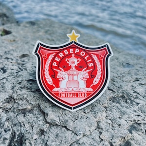 May include: A red, black, and white sticker with the words "PERSEPOLIS FOOTBALL CLUB" and "EST. 1963" in a shield shape.
