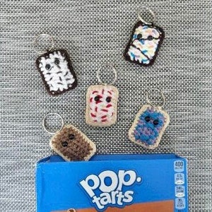 May include: Five crocheted Pop-Tart keychains in different flavors: frosted, brown sugar cinnamon, s'mores, blueberry, and cookies and cream. The keychains are arranged on a gray fabric background with a box of Pop-Tarts Frosted Brown Sugar Cinnamon in the foreground.