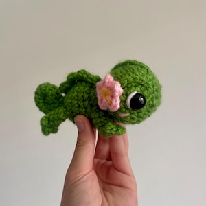 Op de afbeelding: Een handgemaakte groene gehaakte schildpad met een roze bloem. De schildpad heeft grote zwarte ogen en een kleine roze mond. De schildpad wordt in een hand gehouden tegen een effen achtergrond. Het haakwerk is gedetailleerd en de schildpad is klein.