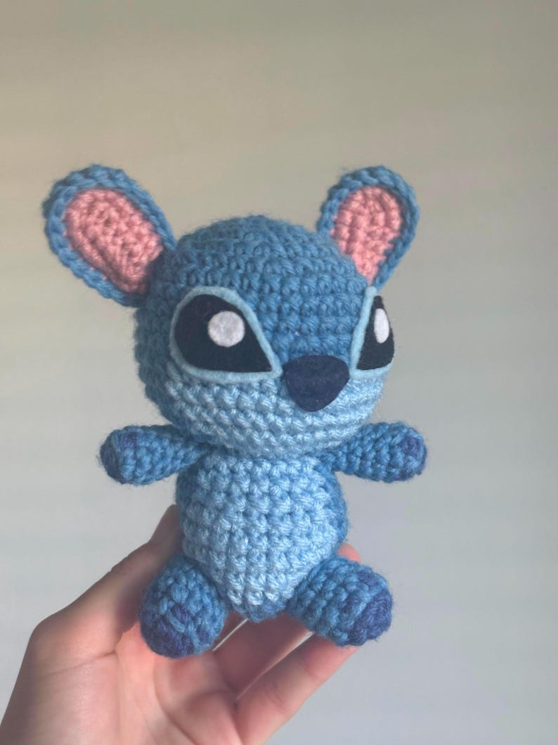 May include: A blue crocheted Stitch plush toy with pink ears and white eyes. The toy is holding its arms out and has a small, black nose.