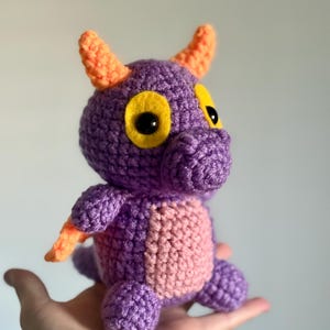 May include: A purple and pink crocheted dragon with orange horns and yellow eyes. The dragon is about 6 inches tall and is made with a soft yarn.