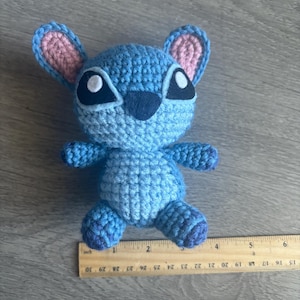 May include: A blue crocheted Stitch plush toy with pink ears and black eyes. The toy is approximately 4 inches tall and is sitting on a wooden ruler.