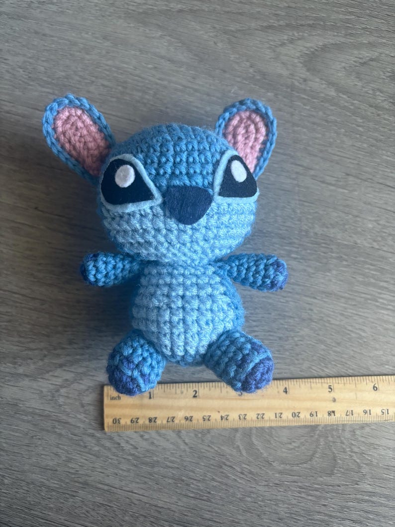 May include: A blue crocheted Stitch plush toy with pink ears and black eyes. The toy is approximately 4 inches tall and is sitting on a wooden ruler.