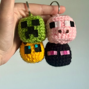 May include: A set of four crocheted Minecraft character keychains. The keychains are green, pink, yellow, and black. Each keychain features a different character from the popular video game.