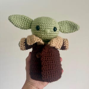 May include: A handmade crochet doll of a character with large ears, black eyes, and a brown coat. The doll is primarily green, with tan and brown accents. The doll has a button closure on the coat. The doll is a Star Wars-themed item.